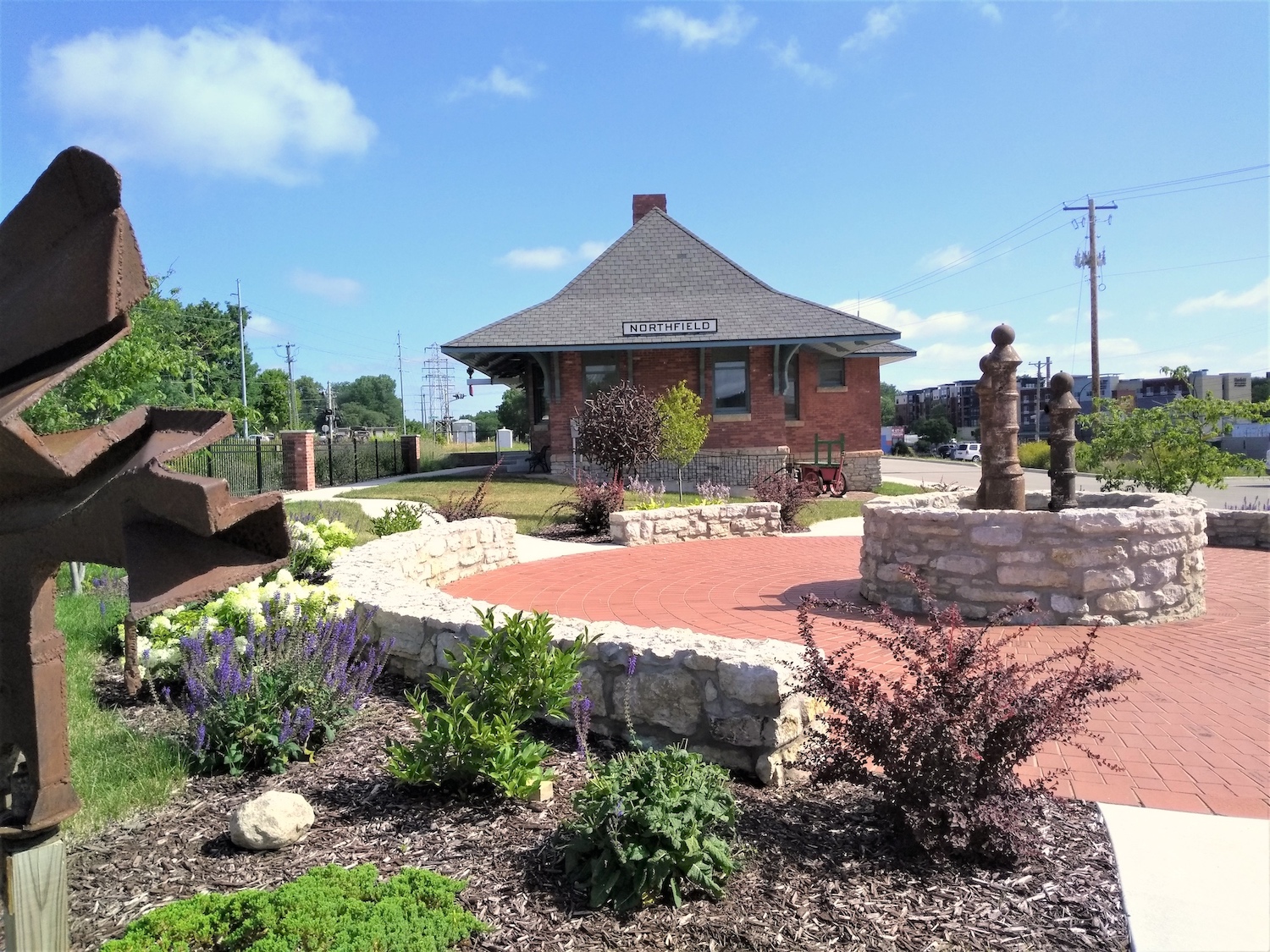 a round brick plaza with a foundation at its center surrounded by a low limestone wall for sitting with the sculpture Wind in the foreground and the sculpture Tree and the depot in the background against a blue sky with wispy clouds